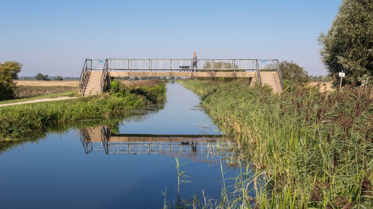 A concrete bridge over a waterway in a flat landscape photographed on a sunny day with blue skies and lush green vegetation on either side of the watercourse, with the bridge reflected in the still, blue water